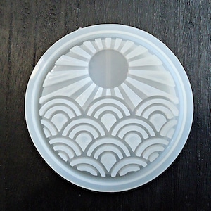 May include: A white silicone mold for resin casting featuring a sun and wave design. The mold is circular and has a raised design of a sun with rays and a wave pattern below.