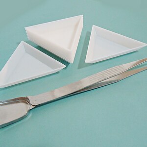 May include: A set of four white triangular trays and a pair of silver tweezers with a small silver scoop. The trays are stacked on top of each other.