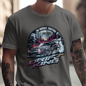 May include: Gray t-shirt featuring a graphic of a drifting car with the text "DRIFT KING" in pink and blue. The design includes a cityscape and the words "THE ORIGINAL MASTERS" in a circular banner.