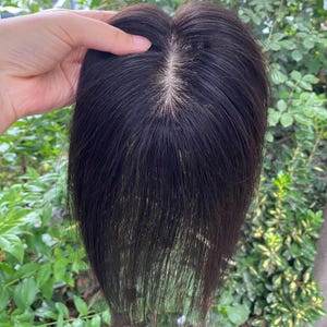 May include: A dark brown hair topper with a natural-looking part. The hairpiece is held up against a backdrop of green foliage, showcasing its color and texture. The hair appears to be smooth and straight.