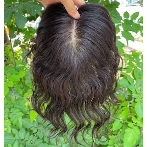 Puede incluir: Una peluca marrón oscuro con cabello ondulado hasta los hombros. La peluca está sujeta por una mano, mostrando el cuero cabelludo y la raya del cabello. El cabello tiene un ligero brillo, y el estilo parece natural y cotidiano.