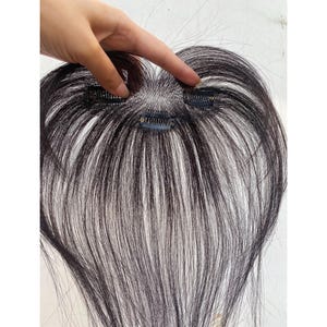 May include: A dark brown hairpiece with three small clips attached. The hairpiece has a natural look with a mix of dark and lighter strands. The hairpiece is designed to add volume or change the hairstyle.