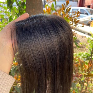 Puede incluir: Una pieza de cabello marrón oscuro con mechones largos y lisos. La pieza de cabello se sostiene, mostrando el color y la textura naturales del cabello. La pieza de cabello parece ser un postizo o una peluca, diseñada para agregar volumen o cobertura.