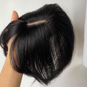May include: A dark black hairpiece with a natural-looking part. The hairpiece appears to be made of real hair and is held in a hand. The hair is styled with a slight wave.