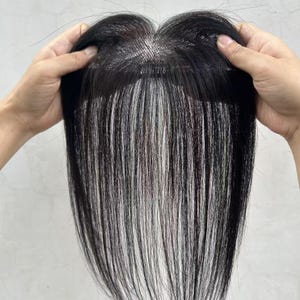 Puede incluir: Una pieza de cabello negra con mechones largos y lisos y una línea de cabello de aspecto natural. La pieza de cabello tiene una mezcla de mechones negros y blancos, que ofrecen un efecto de resaltado. La pieza de cabello está sostenida por dos manos, mostrando su diseño y textura.