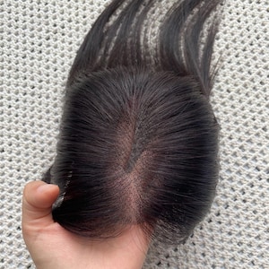 Peut inclure: Un complément capillaire brun foncé doté d'une base en dentelle centrale avec une raie visible. Les cheveux sont lisses et droits, s'étendant depuis la base en maille. La pièce est conçue pour couvrir le sommet du crâne, offrant une apparence naturelle du cuir chevelu à travers le matériau en dentelle fine.