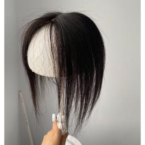 Op de afbeelding: Een donkerbruin haarstuk met lange pony, tentoongesteld op een witte mannequinhoofd. Het haarstuk wordt door een hand vastgehouden en de achtergrond is een effen, lichtgrijze muur. Het haar lijkt steil en glad.