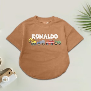 May include: A short-sleeved, terracotta-coloured shirt with the name "RONALDO" in white capital letters above a row of cartoon vehicles. The vehicles include a yellow digger, a blue truck, a grey cement mixer, a red fire engine, and a green tractor.