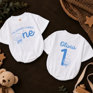 May include: Two white short-sleeved shirts with blue graphics. One shirt features the text "THE SUMMER I TURNED one" with a seashell icon. The other shirt displays the name "Olivia" above the number "1" and a seashell icon with small stars.