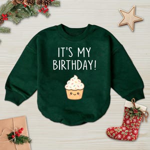 May include: A dark green sweatshirt with the text "IT'S MY BIRTHDAY!" in white. Below the text is a cartoon cupcake with a smiling face and sprinkles. The sweatshirt is a festive item, perfect for a birthday celebration.