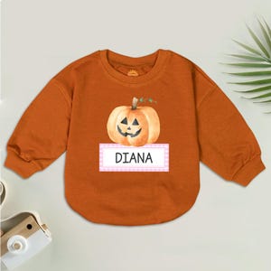 May include: An orange long-sleeved sweatshirt featuring a watercolor pumpkin graphic with a smiling face. The name "DIANA" is printed on a white and pink striped label below the pumpkin. The sweatshirt is a fall-themed item.