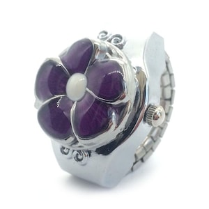May include: Silver-toned ring watch with a purple flower design. The flower has a white center and is set on a stretch band. The watch has a small dial on the side.