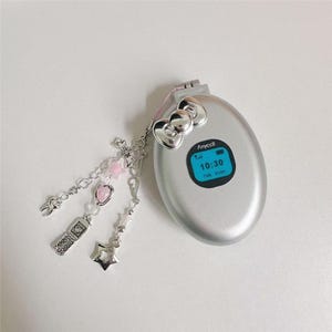 May include: Silver flip phone with a blue screen displaying the time 10:30. The phone has a Hello Kitty bow detail and a charm with a cross, pink beads, a strawberry, a star, and a small phone. The phone is on a white surface.