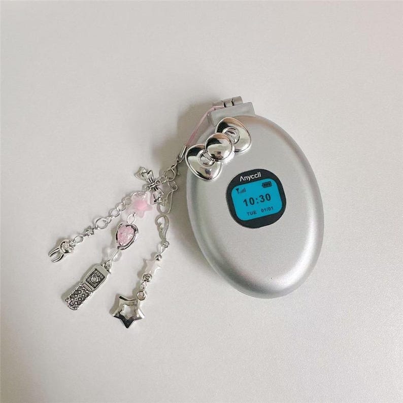 May include: Silver flip phone with a blue screen displaying the time 10:30. The phone has a Hello Kitty bow detail and a charm with a cross, pink beads, a strawberry, a star, and a small phone. The phone is on a white surface.