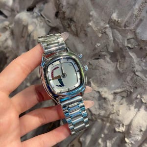 May include: A silver-toned wristwatch with a metallic link band and a square-shaped case. The watch face is white with a stylized, multi-colored graphic element on the left side, featuring red, blue, and brown segments, and simple silver hands.