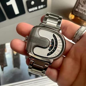 May include: A silver-toned wristwatch with a metallic link band and a unique, asymmetrical case design. The dial features a textured white surface, a curved display window with numerical markings, and the text BESTWIN printed in black lettering.