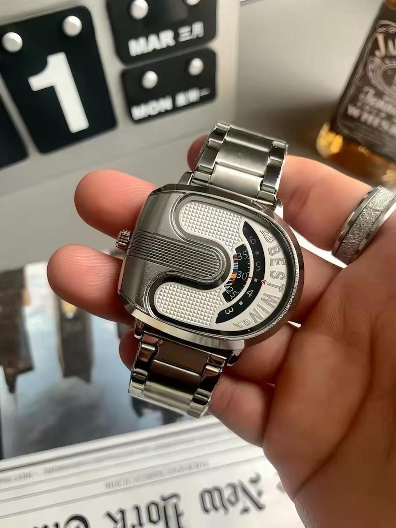 May include: A silver-toned wristwatch with a metallic link band and a unique, asymmetrical case design. The dial features a textured white surface, a curved display window with numerical markings, and the text BESTWIN printed in black lettering.