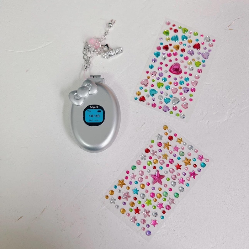 May include: Silver flip phone with a bow-shaped accent and a screen displaying 10:30. Includes a charm with pink beads and a small shoe. Two sheets of colorful gem stickers, one with hearts and the other with stars, are also present.