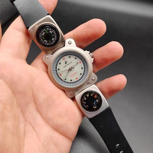 May include: A silver-toned wristwatch featuring a round white dial with the text Remy Martin and Compass, accented by red and black markers. The watch includes two integrated circular compasses on the black rubber strap, one above and one below.