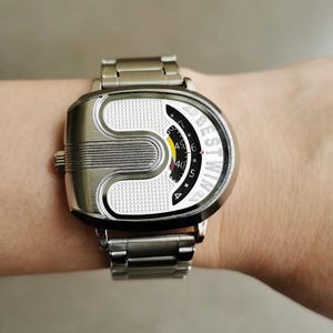 May include: A silver-toned wristwatch with a metallic link band and a unique U-shaped case. The watch face features a textured white surface, a black curved display with the text BESTWIN, and a yellow and red indicator for timekeeping.