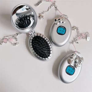 May include: Silver compact mirror and brush set with a matching phone-shaped accessory. The mirror is open, revealing the brush and reflection. The phone-shaped accessories display the time 10:30. Each item is adorned with a decorative chain.