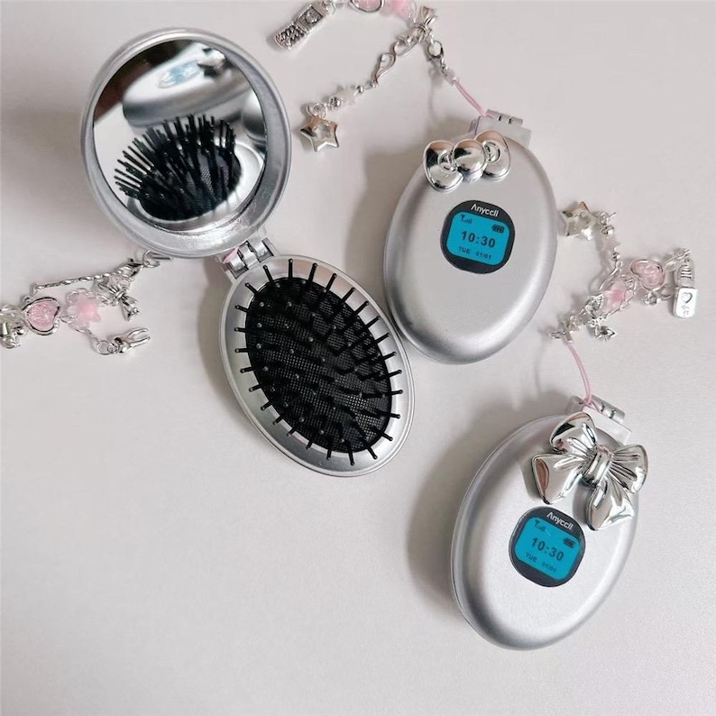 May include: Silver compact mirror and brush set with a matching phone-shaped accessory. The mirror is open, revealing the brush and reflection. The phone-shaped accessories display the time 10:30. Each item is adorned with a decorative chain.