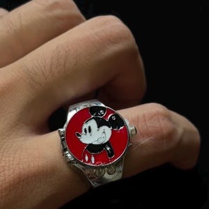 Op de afbeelding: Zilverkleurige ring met een rode wijzerplaat met een lachend Mickey Mouse-ontwerp. De ring heeft een gedetailleerde band met decoratieve elementen. De Mickey Mouse-afbeelding is in zwart, wit en rood.