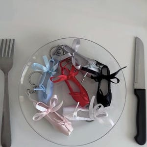 Può includere: Una collezione di portachiavi a forma di scarpette da danza in miniatura nei colori azzurro, rosso, nero, rosa, bianco e argento, ciascuno con un fiocco in nastro di raso, disposti su un piatto di vetro trasparente tra una forchetta e un coltello.