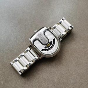 May include: A silver-tone wristwatch with a white and silver link bracelet. The watch face features a unique U-shaped design with a textured silver surface, a black curved display area with yellow accents, and the text BESTWIN printed on the side.