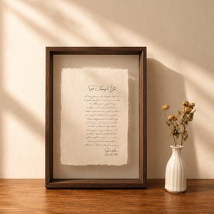 May include: A dark wood frame containing a piece of cream-colored paper with deckled edges and black cursive text titled Still Among You, featuring a signature and date at the bottom, displayed next to a small white vase with dried flowers.