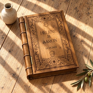 May include: A rectangular wooden box designed to resemble a vintage book with a hinged spine. The cover features intricate laser-engraved floral borders and the text "HELENE & AARON" followed by the date "14.10.2024" centered on the front panel.