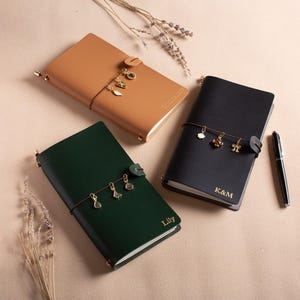 May include: Three leather journals in tan, black, and dark green, each with a button closure and a decorative charm. The black journal has the initials "K&M" and the green journal has the name "Lily" in gold lettering. A black pen is also present.