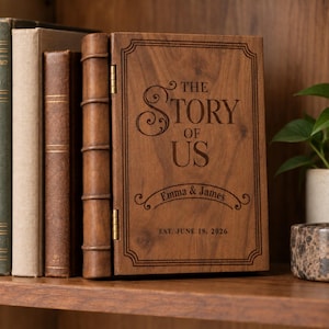 May include: A rectangular wooden box designed to resemble a book with a hinged spine. The front cover is engraved with the text THE STORY OF US, the names Emma & James, and the date EST. JUNE 18, 2026, framed by a decorative border.
