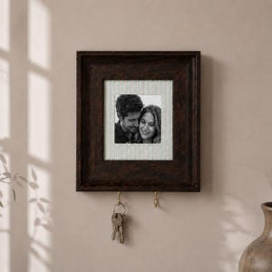 May include: A dark brown wooden wall-mounted photo frame with a square centre cutout displaying a black and white photograph of two people. The frame features two brass-coloured metal hooks at the bottom, with a set of keys hanging from one hook.