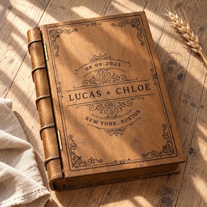 May include: A rectangular wooden box designed to resemble a vintage book with a hinged spine. The cover features laser-engraved decorative scrollwork, the date 08.08.2025, the names LUCAS + CHLOE, and the text NEW YORK, BOSTON centered on the front.