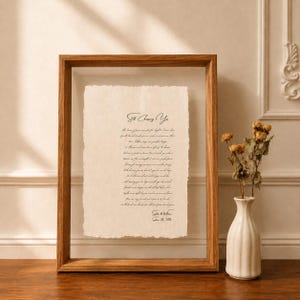 Op de afbeelding: Een rechthoekige houten zwevende lijst met een vel crèmekleurig papier met gescheurde randen. Het papier bevat zwarte cursieve tekst met de titel Still Choosing You, gevolgd door een gedicht, de namen Sophie & William en de datum 28 juni 2026.