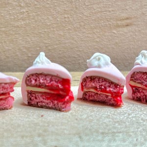 May include: Four miniature cake slices with pink icing, red filling, and white whipped cream topping. The cake slices are arranged in a row on a light brown surface. The cake has a layered design.
