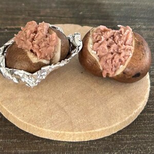 May include: Two baked potatoes, each filled with a pinkish-brown topping, sit on a round, light brown wooden board. One potato is wrapped in aluminum foil. The potatoes are on a dark wooden surface.