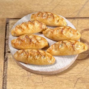 May include: Six miniature, golden-brown pastries are arranged on a small, round, light-coloured wooden surface. The pastries have a textured, baked appearance, with visible details and a glossy finish. The background is a wooden surface.