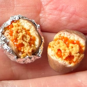 Könnte beinhalten: Zwei Miniatur-Lebensmittel, eines in Folie gewickelt, das andere nicht. Beide haben eine hellbraune Außenseite und sind mit einer gelb-orangen Mischung gefüllt, die möglicherweise ein Miniatur-Sandwich oder ein ähnliches Lebensmittel darstellt.