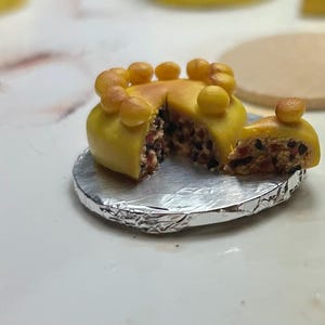 Può includere: Una torta gialla in miniatura con una fetta tagliata, che rivela un interno scuro e macchiato. La torta è decorata con piccoli accenti gialli rotondi e poggia su una base di carta stagnola argentata. È un alimento in miniatura.