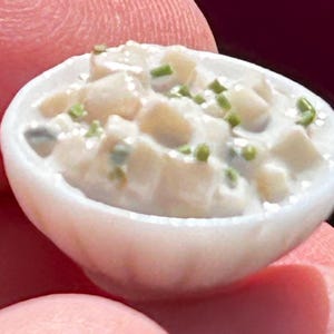 May include: A miniature white bowl filled with a creamy, off-white dish, garnished with small green pieces. The food appears to be a potato salad or similar side dish. The bowl is held between fingers.