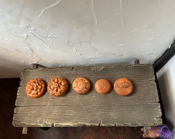 May include: Five round, brown, ceramic tiles with different designs. The tiles are arranged in a row on a wooden surface.