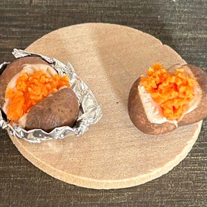 Könnte beinhalten: Zwei Ofenkartoffeln, eine in Folie, beide mit orangenem Belag, liegen auf einer runden, beigen Oberfläche. Die Kartoffeln haben eine braune Schale und ein weißes Inneres. Der orangefarbene Belag ist fein gehackt.