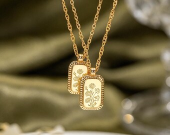 Könnte beinhalten: Zwei goldfarbene Halsketten mit rechteckigen Anhängern mit abgerundeten Ecken und gewelltem Rand. Jeder Anhänger ist mit einem zarten Blumenmuster graviert. Die Ketten hängen an dünnen, gedrehten goldfarbenen Metallketten.