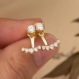 May include: Gold-coloured earrings featuring two large, clear, round gemstones at the top and a row of smaller, clear stones along the bottom. The earrings are held in a hand against a neutral background.