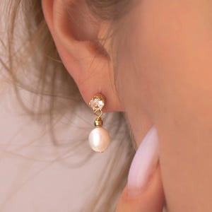May include: Gold-toned earrings featuring a small, clear gemstone at the top and a dangling white pearl. The earrings are worn on an ear, showcasing the design and elegance of the jewelry. The pearl is a classic, timeless design.