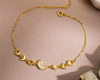 May include: Gold-toned bracelet featuring a crescent moon and circular charms with white accents. The delicate chain is displayed on a pink surface, with a sprig of greenery in the lower right corner.
