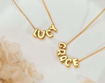 Könnte beinhalten: Zwei goldfarbene Halsketten mit einzelnen Buchstabenanhängern. Eine Kette bildet den Namen LUCY und die andere den Namen GRACE. Die Halsketten haben feine, zarte Goldketten und die Buchstaben haben eine glatte, abgerundete Metalloberfläche.