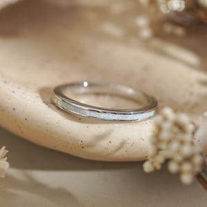 May include: A silver ring with a band of white opal. The ring is set on a beige, textured surface, with dried flowers in the background. The ring is a delicate piece of jewelry.
